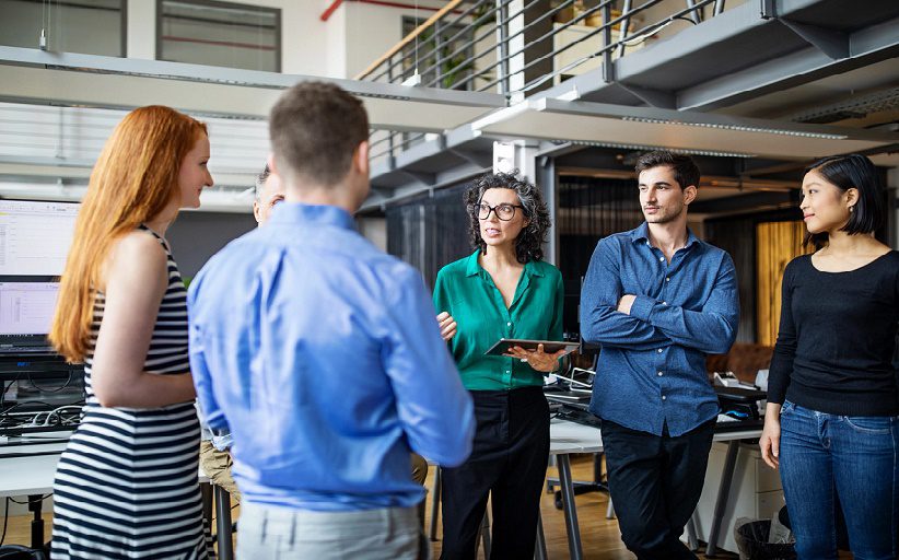 Un grupo de ejecutivos, conformado por dos hombres y tres mujeres, discuten de pie en medio de una sala de juntas.