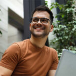 Smiling man with laptop sitting outdoors.